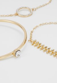 Ensemble de bijoux dorés comprenant un bracelet lisse avec une pierre transparente, une chaîne en maillons circulaires et une chaîne avec des pierres transparentes alternées.