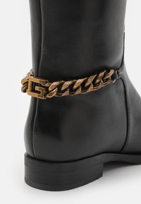 Botas de cuero negro con un acento de cadena de bronce gruesa con un logo. Textura suave, puntera redonda y tacón bajo apilado.