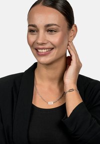 Silver chain necklace and bracelet with rectangular accents on a model wearing a black top and jacket. Smooth metal finish.