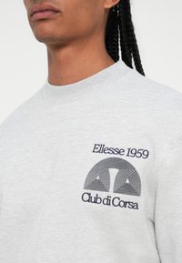 Sivá mikina s okrúhlym výstrihom. Má na hrudi námornícky vyšívaný grafik s textom "Ellesse 1959" a "Club di Corsa".