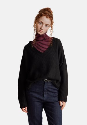 Donna con capelli ricci rossi che indossa un maglione nero oversize con scollo a V sopra un dolcevita a righe bordeaux e jeans scuri con cintura nera.