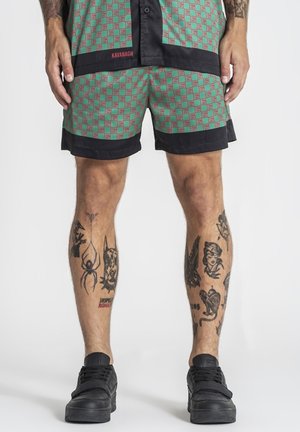 Muž nosiaci zeleno-červeno kockované šortky a zodpovedajúcu košeľu s čiernym lemovaním, čierne tenisky a rôzne tetovania na oboch nohách.