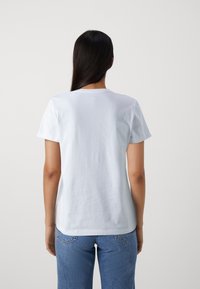 Camiseta blanca de algodón con mangas cortas, cuello redondo, textura suave y un corte relajado, vista desde la espalda. Combinada con vaqueros azules.