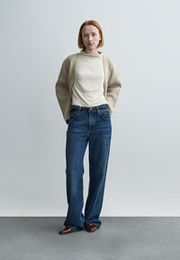 Νεαρή γυναίκα με κοντά κόκκινα μαλλιά, ντυμένη με μπεζ ζακέτα, κρεμ τοπ, φαρδιά μπλε τζιν και καφέ παπούτσια, στεκόμενη μπροστά σε ανοιχτό γκρι τοίχο.