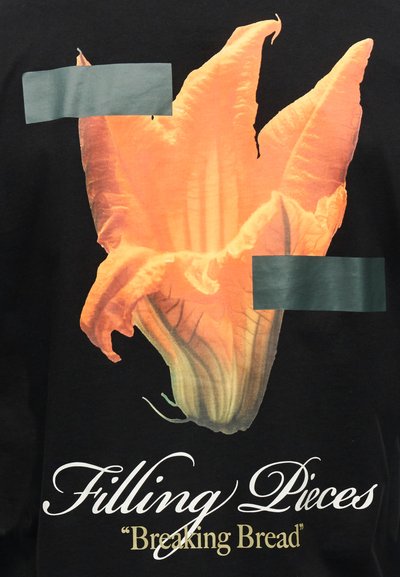 Λαμπερό πορτοκαλί λουλούδι σε μαύρο φόντο με το κείμενο "Filling Pieces" και "Breaking Bread" από κάτω.