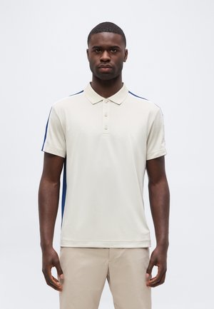 Homme portant un polo beige à manches courtes avec des rayures bleu marine sur les épaules et un pantalon beige clair, se tenant devant un fond blanc.