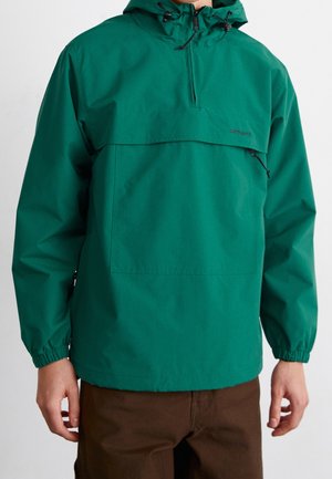 Homme portant une veste à capuche verte avec fermeture éclair frontale et poignets élastiques, associée à un pantalon marron, sur un fond uni.