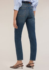 Jeansy z wysokim stanem w ciemnoniebieskim kolorze, o prostych nogawkach, z pięcioma kieszeniami, subtelną teksturą i ciemnym szwem; w zestawie z obcasami typu mule.