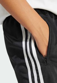 Černé sportovní šortky s bílými pruhy, mají boční kapsu na zip a hladkou strukturu, detail ruky v kapse.