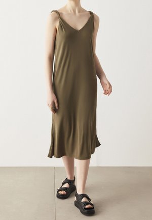 Robe midi sans manches de couleur vert olive, avec un décolleté en V, dotée de bretelles nouées et de fentes sur les côtés. Associée à des sandales plateforme noires.