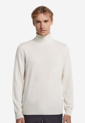Ung mand iført en simpel hvid rullekrave og sorte bukser, stående mod en hvid baggrund med et neutralt udtryk.