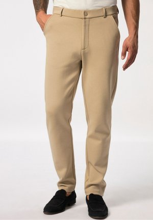 Man wearing beige slim-fit trousers with side pockets and black loafers, standing against a plain light background.