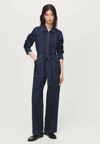 Combinaison en denim avec zip sur le devant, col et jambes larges ; dispose d'une ceinture en tissu et de coutures contrastantes. Couleur : bleu foncé.