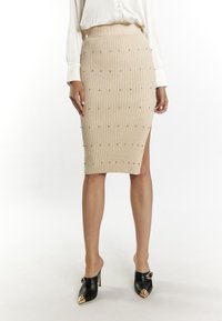 Beige ribgebreide pencil skirt met een zijsplits, versierd met kleine zilveren studs, gecombineerd met zwarte puntige hakken.