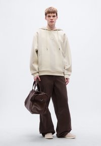 Μπεζ πλεκτό φούτερ με κουκούλα και κεντρικό έμβλημα, συνδυασμένο με καφέ παντελόνι φαρδιάς γραμμής. Το μοντέλο κρατάει μια σκούρα καφέ δερμάτινη τσάντα με ασημί εξαρτήματα.