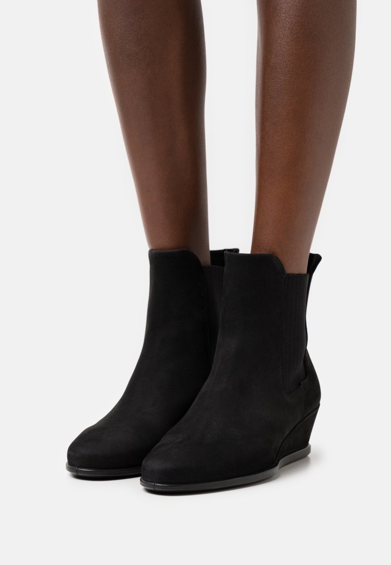 Bottines en daim noir à talons compensés portées sur des jambes à la peau foncée, présentées sur un fond blanc uni.