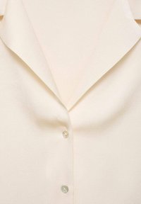 Cream blouse with a soft drape, featuring a point collar and three buttons in a pearlescent finish, highlighting a smooth, textured fabric.