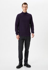 Paarse gebreide coltrui met ribbeltextuur, gecombineerd met donkere jeans en zwarte enkellaarsjes, staande op een effen achtergrond.