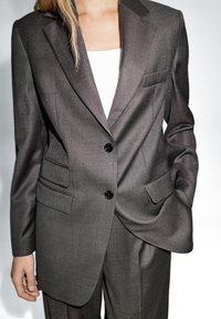 Gray tailored blazer made of smooth fabric, featuring a notched lapel, two front pockets, and two black buttons. Structured fit with clean lines.