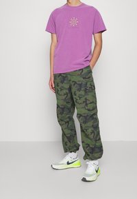 Violetti t-paita rintakehän graafisella kuviolla, yhdistettynä vihreisiin maastokuvioisiin cargo-housuihin ja vaaleanharmaisiin Nike-tossuihin, joissa on neonkoristeet.