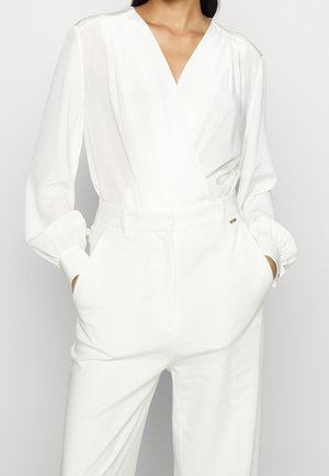 Vrouw draagt een wit langärmelig V-hals blouse, ingestopt in hooggehuisde witte broek, met handen in de zakken.