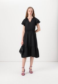 Black dress with short ruffled sleeves, V-neck, cinched waist, and tiered hem. Paired with purple strappy sandals and a black bag.