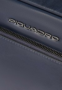 Borsellino in pelle blu navy con logo "Piquadro" argentato, cerniera metallica e superficie liscia con dettagli di cucitura visibili.