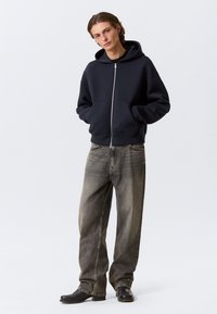 Sweat à capuche zippé bleu marine avec une large capuche et des poches avant, associé à un jean gris ample et des chaussures noires. Texture de tissu lisse.