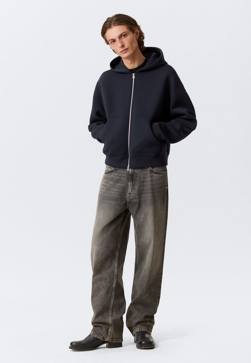 Sweat à capuche zippé bleu marine avec une large capuche et des poches avant, associé à un jean gris ample et des chaussures noires. Texture de tissu lisse.