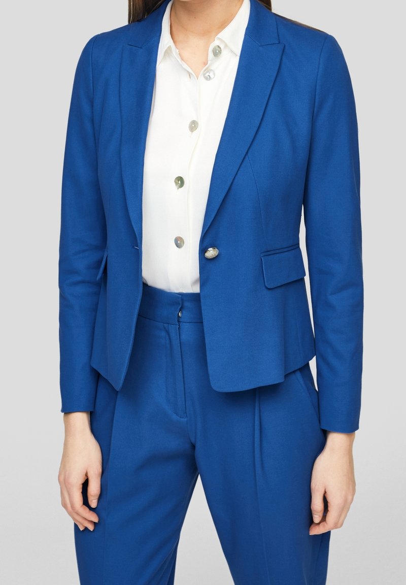 Blå skräddarsydd kavaj med en enkel knapp, spetsiga slag och framfickor, bar över en vit skjorta med flera knappar och en slät yta.