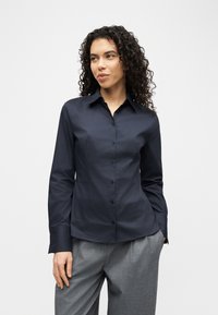 Γυναίκα με σγουρά σκούρα μαλλιά, ντυμένη με στενή μπλε ναυτική πουκάμισο με κουμπιά και άνετο γκρι παντελόνι, στέκεται μπροστά σε ένα απλό λευκό φόντο.