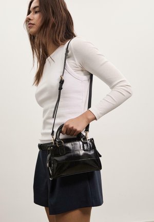 Sac à main en faux cuir noir avec un motif crocodile, deux poignées, une bandoulière et des accents en métal doré. Porté par une personne.
