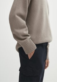 Beżowy sweter robiony na drutach z ściągaczami, w połączeniu z ciemnoniebieskimi spodniami cargo z bocznymi kieszeniami. Miękka faktura, casualowy design.