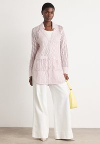 Lichtroze gebreide cardigan met open weefdesign, lange mouwen, twee voorzakken en knoopsluiting. Gecombineerd met witte hoge brede broek.