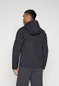 Wasserdichte schwarze Jacke mit Kapuze, mit glatter Textur, seitlichen Reißverschlusstaschen und verstellbaren Bündchen. Rückansicht, getragen über grauen Hosen.