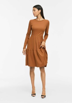Femme en robe marron rouille à manches longues et longueur genoux, tenant la jupe, regardant vers sa gauche, portant des talons noirs ouverts à l’avant.