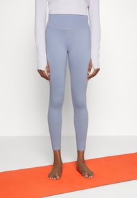 Leggings azzurri chiari realizzati in un materiale elasticizzato, con una vita alta e una consistenza liscia, indossati a piedi nudi su un tappetino yoga arancione.