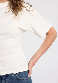 Cream-colored cotton t-shirt with short sleeves, slightly gathered at the side, paired with denim jeans. Textured fabric, casual fit.