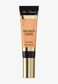 Fond de teint Soft Matte Born This Way de Too Faced dans un tube pêche, avec des accents dorés et un capuchon noir. Texte : "Tenue 24 heures" et "Acide hyaluronique."