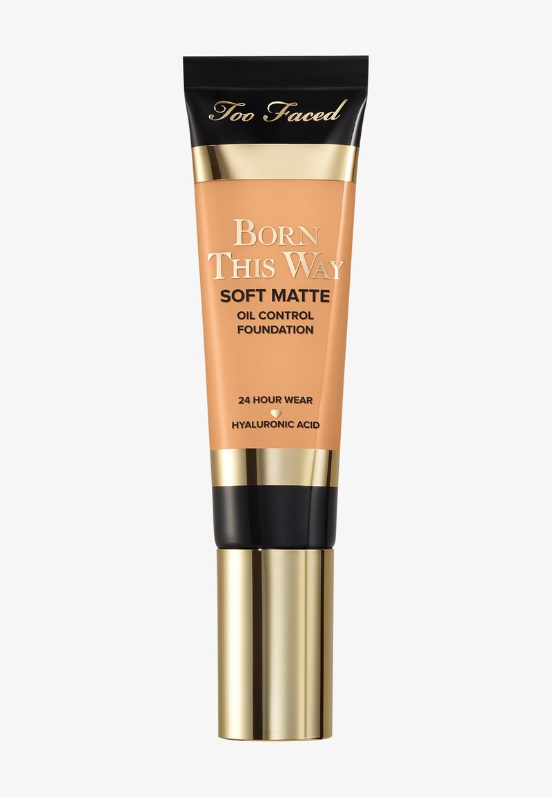 Fond de teint Soft Matte Born This Way de Too Faced dans un tube pêche, avec des accents dorés et un capuchon noir. Texte : "Tenue 24 heures" et "Acide hyaluronique."