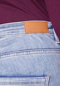 Jeans de mezclilla de color azul claro con una parche de cuero marrón etiquetado "VERO MODA". La tela tiene una textura ligeramente desgastada.