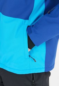 Giacca blu e turchese con tasca con zip e dettagli in velcro. Il materiale è morbido e leggero, con un design a blocchi di colore.