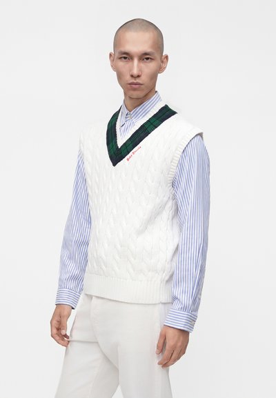 Homme portant un gilet sans manches blanc en maille torsadée avec un col en V vert et noir par-dessus une chemise à rayures bleues et blanches à boutons.