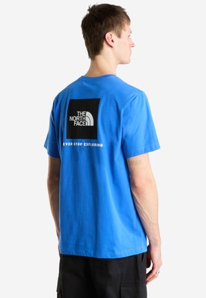 Joven de perfil lleva una camiseta azul con el logo "The North Face" y el texto "Never Stop Exploring" en la espalda, combinada con pantalones negros.