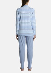 Donna in piedi a piedi nudi che indossa un top a maniche lunghe azzurro chiaro con un motivo e pantaloni azzurri chiari uniformi, vista da dietro contro uno sfondo bianco.