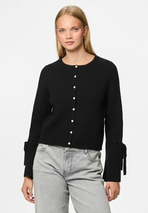 Svart stickad cardigan med rund halsringning, knappstängning fram och betonar ärmar med band. Texturerad tyg och pärlknappdetaljer.