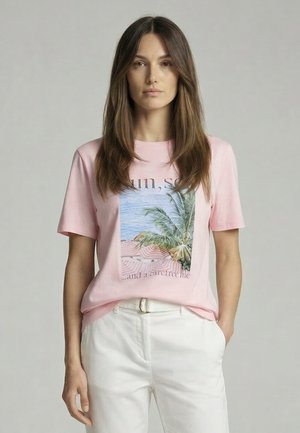 Woman with long brown hair wearing a pink T-shirt with a beach and palm tree print, paired with white pants, hands in pockets, neutral background.