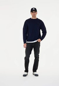 Mornarsko modra kapuca, bela majica, črn jeans, in bele superge z modrimi detajli. Oseba nosi tudi črno kapo.