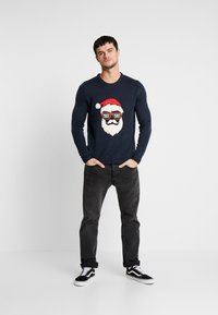 Marineblauwe trui met een Santa-graphic met zonnebril, witte baard en rode hoed. Gecombineerd met donkere jeans en zwarte sneakers.