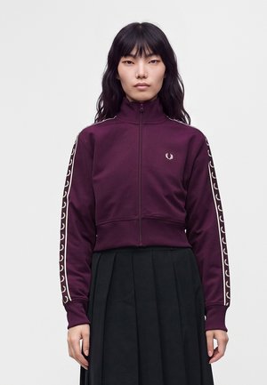 Femme aux longs cheveux noirs porte une veste de sport violette zippée avec des bandes blanches de logo sur les manches et une jupe plissée noire sur un fond uni.